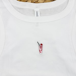 May include: White vest top featuring a small embroidered cocktail design. The cocktail is red and white with a black straw. The top is made of a soft material.