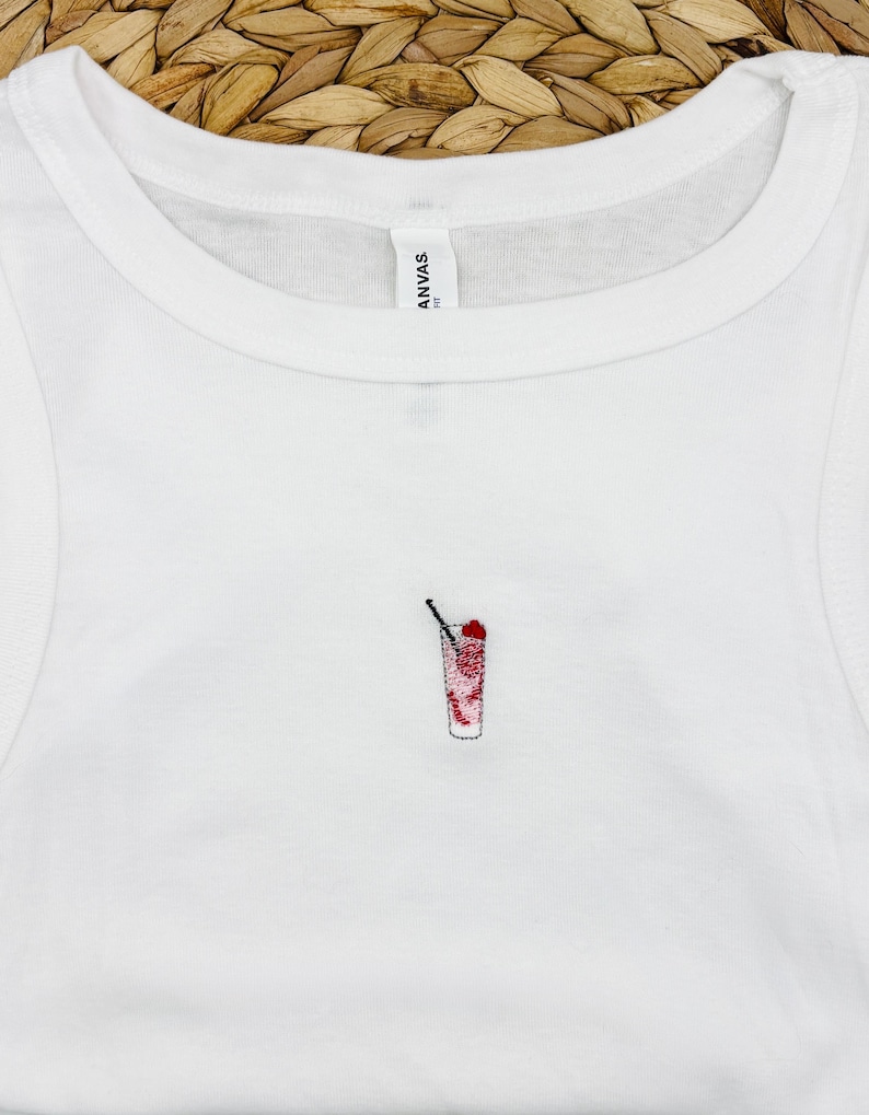 May include: White vest top featuring a small embroidered cocktail design. The cocktail is red and white with a black straw. The top is made of a soft material.