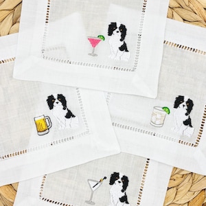 May include: Set of four white linen cocktail napkins. Each napkin features an embroidered black and white Cavalier King Charles Spaniel with a different cocktail: a martini, margarita, beer, and a cocktail with ice and lime.