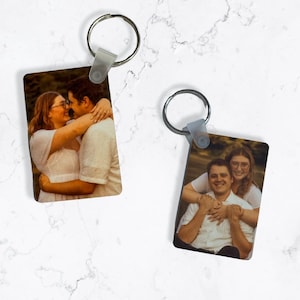 May include: Two rectangular keyrings with metal rings. Each keyring features a colour photograph of a couple embracing. The keyrings are on a white marble surface.