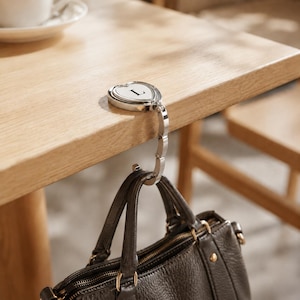 May include: A silver-toned metal bag hook with a heart-shaped top featuring the letter L in a black serif font. The hook is secured to the edge of a table, supporting a dark brown textured leather handbag by its two handles from the bottom hook.