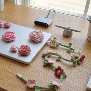 May include: A collection of handmade crochet flowers in varying shades of pink, arranged on a silver laptop. A garland of crochet flowers and leaves is also present. The items are on a wooden desk with a notebook, pen, and other accessories.