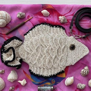 Op de afbeelding: Een beige gehaakte visvormige portemonnee met zwarte rand en een decoratief knoopoog. De portemonnee ligt op een roze bloemen sjaal, omringd door schelpen en een zwarte riem. Er is ook een haaknaald aanwezig.