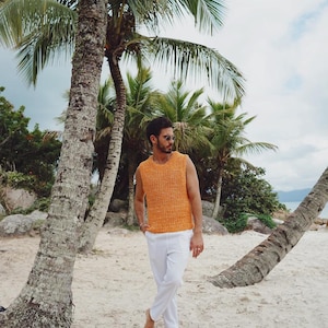 Pu&ograve; includere: Un maglione in maglia senza maniche di colore arancione brillante con scollo rotondo e consistenza spessa, abbinato a pantaloni bianchi semplici. Il capo presenta una trama larga e traspirante con un design essenziale e pulito.