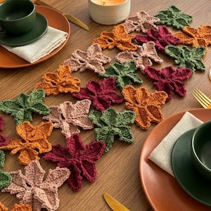 May include: A table runner composed of interconnected crocheted leaf motifs in shades of deep red, burnt orange, sage green, and dusty pink. The runner features a repeating pattern of textured, multi-lobed leaves joined together in a long strip.