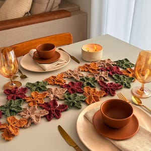 May include: A table runner composed of interconnected crocheted leaves in shades of orange, burgundy, olive green, and beige. The runner is centered on a table set with two beige plates, two terracotta-colored bowls, gold cutlery, and amber glasses.