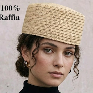 May include: A beige raffia hat with a rectangular crown. The hat is made of woven raffia and has a structured shape. The text "100% Raffia" is in the upper left corner. The hat is worn by a person with curly brown hair and gold hoop earrings.
