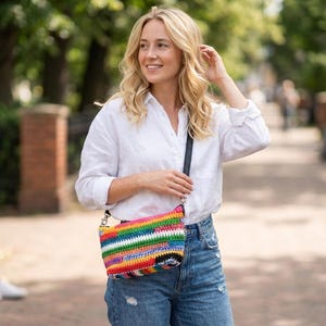 May include: A vibrant, crocheted crossbody bag with a rainbow design. The bag has a black strap and zip, and is worn with a white shirt and blue jeans. The bag is approximately 25 cm wide.