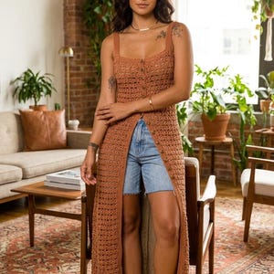 May include: A long, button-down, crochet dress in a warm brown hue. The dress features a square neckline and a high slit, paired with denim shorts. The outfit is completed with blue sandals. The setting is a well-lit living room with plants and a patterned rug.