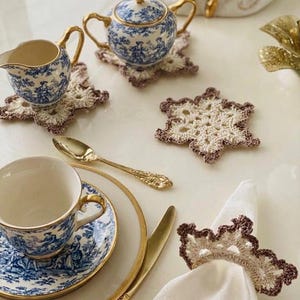May include: A collection of cream and brown crocheted coasters and a napkin ring featuring a star-like pattern with scalloped edges. The set is displayed alongside blue and white porcelain tea service pieces, gold-toned cutlery, and a white napkin.