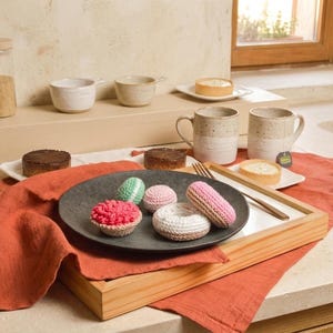 May include: A collection of small, handcrafted crochet food items including a pink cupcake, a green and white macaron, a small pink round piece, and a white and tan donut, all arranged on a dark gray circular plate atop a wooden serving tray.