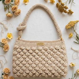 Può includere: Borsa a mano all'uncinetto beige con un motivo a bolle strutturato. La borsa ha un manico annodato e finiture color oro. L'etichetta recita "Bela Crochet". Fiori gialli circondano la borsa.