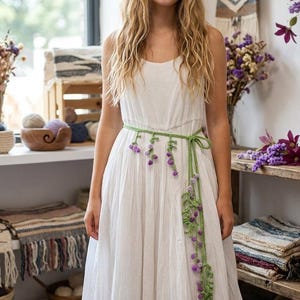 Könnte beinhalten: Ein weißes ärmelloses Maxikleid mit gestuftem Rock und Rundhalsausschnitt, akzentuiert durch einen langen, dünnen grünen Häkelgürtel. Der Gürtel ist mit kleinen, herabhängenden violetten Blumenbüscheln verziert, die vorne am Kleid herabfallen.