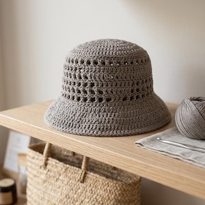 May include: A gray crocheted sun hat with a wide brim sits on a wooden shelf. Two balls of yarn and a crochet hook are next to the hat. A woven basket is below the shelf.