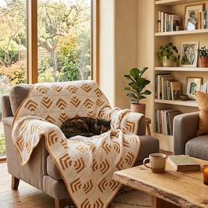 May include: A cozy living room scene featuring a tan armchair draped with a cream and brown geometric patterned blanket. A cat is curled up on the blanket. A wooden coffee table holds candles and a mug. A bookshelf and window are in the background.