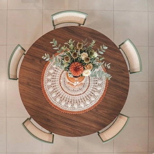May include: A round dark wood dining table surrounded by four chairs with light wood frames and woven cane backrests. A white and rust-colored circular crochet doily sits in the center under a glass vase filled with a bouquet of flowers.
