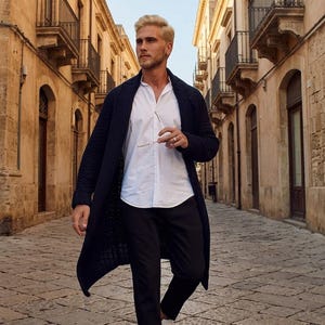 Könnte beinhalten: Eine lange, dunkelblaue Strickjacke über einem weißen Hemd mit Knopfleiste, kombiniert mit einer schwarzen Hose und sauberen weißen Ledersneakern. Das Outfit wird durch eine dünne Brille ergänzt, die beim Gehen in der Hand gehalten wird.