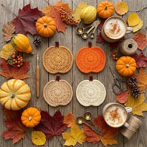 May include: Four round crochet pumpkin coasters in mustard, orange, beige, and cream, arranged on a wooden surface with autumn leaves, small pumpkins, two mugs of coffee, a wooden crochet hook, metal measuring spoons, and twine.