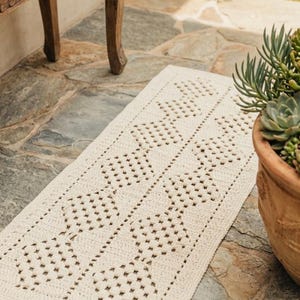 May include: Cream-colored, rectangular crochet rug with a geometric pattern. The rug is placed on a stone surface near a wooden table leg and a terracotta pot with greenery. The rug's design features openwork squares and diamond shapes.