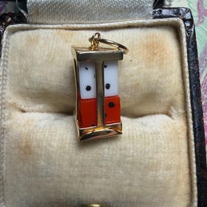 Könnte beinhalten: Ein goldfarbener Anhänger in Form eines Miniatur-Würfelturms mit roten und weißen Würfeln. Der Anhänger hängt an einem goldenen Ring und wird in einer Vintage-Schmuckschatulle mit beigem Samtinterieur präsentiert. Die Würfel haben schwarze Punkte.