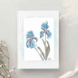 May include: A white journal featuring a watercolor painting of two blue iris flowers with orange accents. The artwork is set against a light blue scalloped border. The journal's cover is plain white.