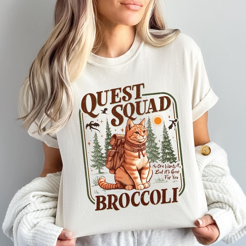 K&ouml;nnte beinhalten: Cremefarbenes T-Shirt mit einer Cartoon-Katze mit Rucksack vor einer Waldkulisse. Der Text lautet "QUEST SQUAD BROCCOLI" mit dem Satz "No One Wants It, But It's Good For You".