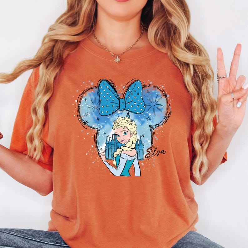 May include: Orange t-shirt featuring a graphic of Elsa from Frozen inside a Mickey Mouse ear silhouette. The design includes a blue bow with white polka dots, a castle, and the name "Elsa" in cursive. The shirt is a solid orange color.