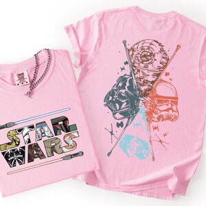 K&ouml;nnte beinhalten: Zwei rosa T-Shirts mit Star Wars-Grafiken. Eines zeigt den Text "STAR WARS" mit Charakterillustrationen und Lichtschwertern. Das andere zeigt Darth Vader, einen Sturmtruppler und den Todesstern. Eine silberne Kette befindet sich auf einem T-Shirt.