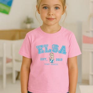 May include: A pink t-shirt featuring the name "ELSA" in light blue block letters. Below the name is "EST. 2013" and a graphic of Elsa from Disney's Frozen. The words "DISNEY PRINCESS" are printed below the graphic.