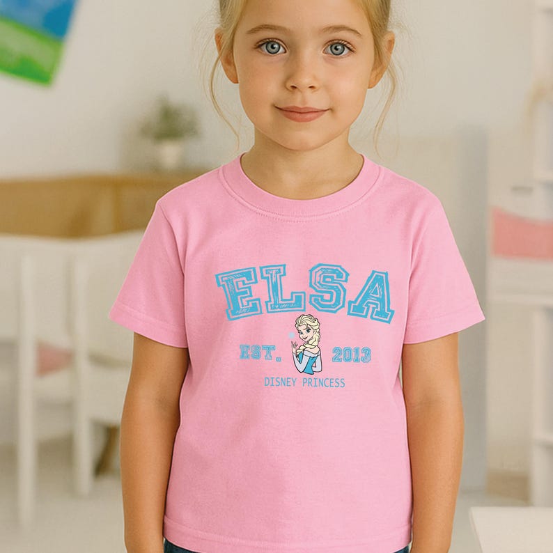 May include: A pink t-shirt featuring the name "ELSA" in light blue block letters. Below the name is "EST. 2013" and a graphic of Elsa from Disney's Frozen. The words "DISNEY PRINCESS" are printed below the graphic.