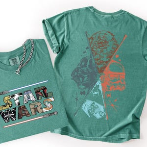 K&ouml;nnte beinhalten: Zwei tealfarbene T-Shirts. Eines gefaltet mit den Worten "STAR WARS" in einer bunten Grafik. Das andere Shirt hat eine Grafik von Darth Vader, einem Sturmtruppler und dem Todesstern. Eine silberne Halskette befindet sich auf dem gefalteten Shirt.