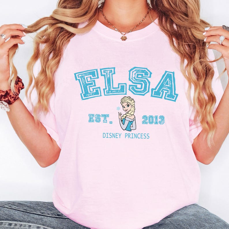 May include: A light pink t-shirt featuring the name "ELSA" in large, light blue block letters. Below the name is "EST. 2013" and a graphic of Elsa, a Disney princess. The words "DISNEY PRINCESS" are printed below the graphic.
