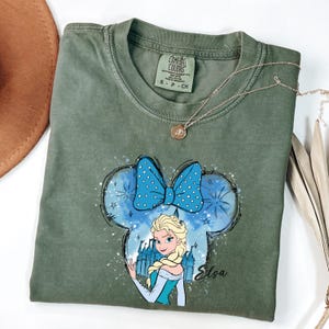 May include: Olive green t-shirt featuring a graphic of Elsa from Frozen inside a Mickey Mouse ear outline with a blue polka dot bow. The word "Elsa" is written in cursive. A gold necklace is draped across the shirt.