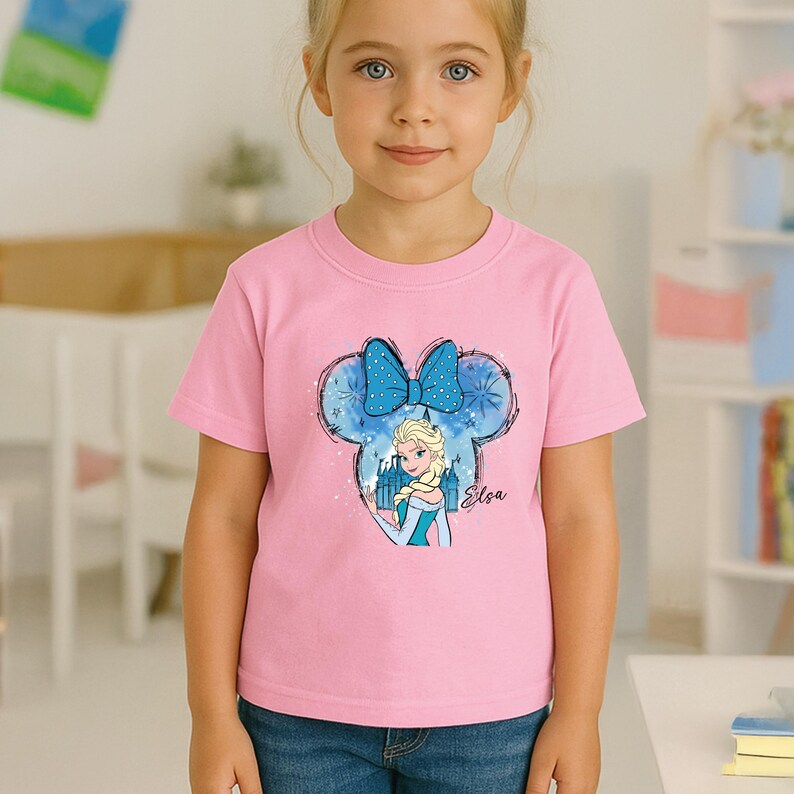 May include: Pink short-sleeved t-shirt featuring a graphic of Elsa from Frozen. The design includes a blue bow, castle silhouette, and the name "Elsa" in script. The shirt is made of a soft material.