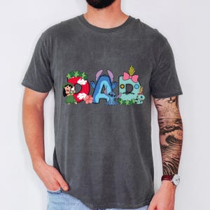 May include: A charcoal gray short-sleeve t-shirt featuring a graphic print that spells the word DAD. The letters are decorated with Lilo and Stitch characters, including Lilo, Stitch, and Scrump, along with tropical flowers and green leaves.