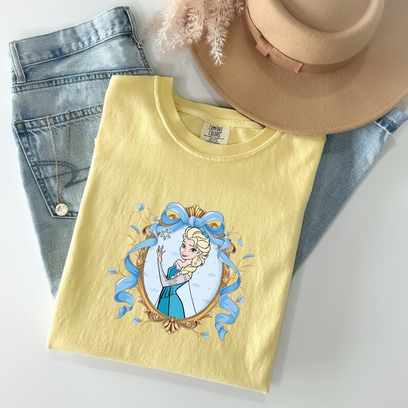 May include: A pale yellow t-shirt featuring a graphic of a blonde character in a blue dress inside an ornate gold oval frame, accented with a large blue ribbon and small snowflakes. The shirt has a Comfort Colors label at the inner neckline.