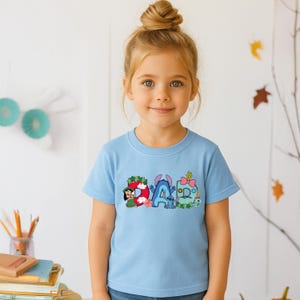 May include: A light blue short-sleeve t-shirt featuring a graphic print of the word DAD. The letter D is decorated with Lilo, the letter A features Stitch, and the letter D includes a pink bow and green foliage elements in a colorful design.