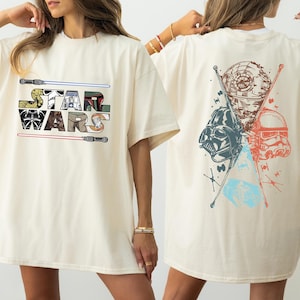 K&ouml;nnte beinhalten: Cremefarbenes T-Shirt mit dem Star Wars-Logo auf der Vorderseite, flankiert von Lichtschwertern. Auf der R&uuml;ckseite sind Darth Vader, ein Sturmtruppler und der Todesstern mit gekreuzten Lichtschwertern abgebildet.