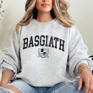 以下が含まれることがあります： 胸に「BASGIATH」の文字が太字の黒色でプリントされたライトグレーのスウェットシャツ。その下に「WAR COLLEGE」の文字が入った紋章があります。スウェットシャツはカジュアルなスタイルです。