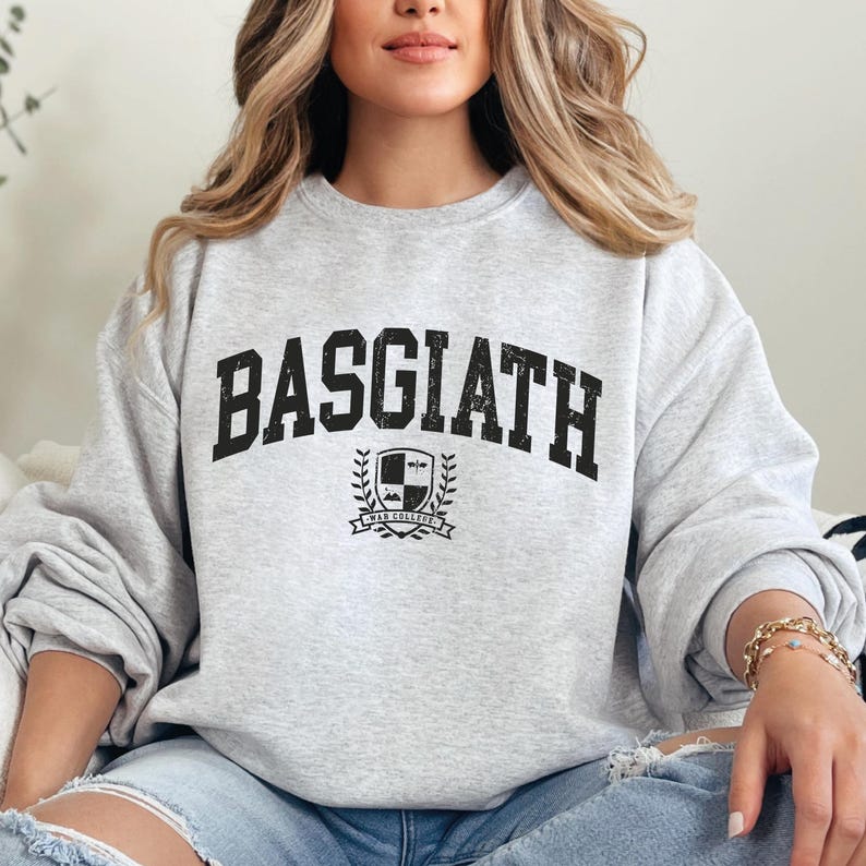 以下が含まれることがあります： 胸に「BASGIATH」の文字が太字の黒色でプリントされたライトグレーのスウェットシャツ。その下に「WAR COLLEGE」の文字が入った紋章があります。スウェットシャツはカジュアルなスタイルです。