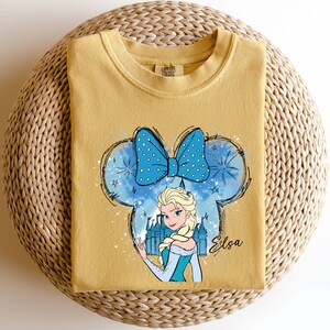May include: Mustard yellow t-shirt featuring a cartoon illustration of Elsa from Frozen. The design includes a blue bow with white polka dots, a castle, and the name "Elsa" in cursive. The shirt is displayed on a woven, circular surface.