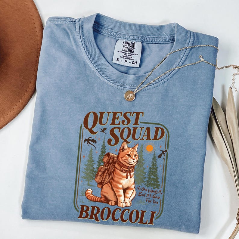 K&ouml;nnte beinhalten: Hellblaues Comfort Colors T-Shirt mit einer Grafik einer orangefarbenen Tabby-Katze mit Rucksack, den Worten "QUEST SQUAD BROCCOLI" und dem Text "No One Wants It, But It's Good For You". Eine goldene Halskette liegt auf dem Shirt.