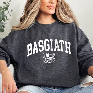 以下が含まれることがあります： 胸に「BASGIATH」の文字が白くかすれた文字でプリントされたダークグレーのスウェットシャツ。その下に「BAR COLLEGE」の文字が入った紋章があります。クルーネックのスタイルのスウェットシャツです。