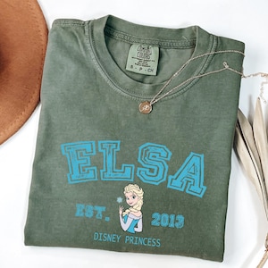 May include: An olive green t-shirt with the name "ELSA" in blue block letters. Below the name is a graphic of Elsa from Disney's Frozen, the text "EST. 2013" and "DISNEY PRINCESS". A gold necklace with a pendant is on the shirt.