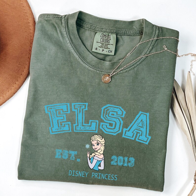 May include: An olive green t-shirt with the name "ELSA" in blue block letters. Below the name is a graphic of Elsa from Disney's Frozen, the text "EST. 2013" and "DISNEY PRINCESS". A gold necklace with a pendant is on the shirt.