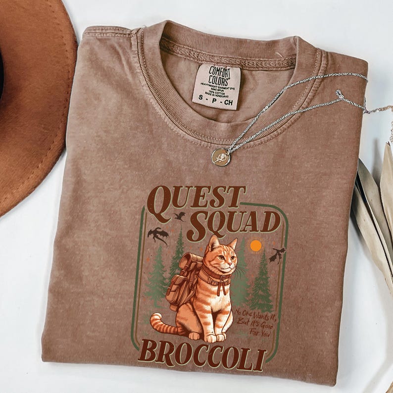 K&ouml;nnte beinhalten: Ein braunes T-Shirt mit einer Grafik, die eine Katze mit Rucksack, die Worte "Quest Squad" und "Broccoli" zeigt. Das Shirt hat einen Rundhalsausschnitt und kurze &Auml;rmel. Eine silberne Halskette mit Anh&auml;nger liegt auf dem Shirt.