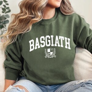 以下が含まれることがあります： 胸に白の擦り切れた文字で「BASGIATH」と書かれたオリーブグリーンのスウェットシャツ。その下に、盾、本、バナーが付いた紋章のデザインがあります。スウェットシャツはクルーネックと長袖です。