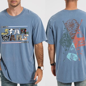 K&ouml;nnte beinhalten: Blaues T-Shirt mit dem Schriftzug "STAR WARS" in einer farbenfrohen, stilisierten Schriftart mit Lichtschwertern. Auf der R&uuml;ckseite des Shirts befindet sich eine Grafik von Star Wars-Charakteren und -Symbolen in Blau-, Orange- und Gr&uuml;nt&ouml;nen.