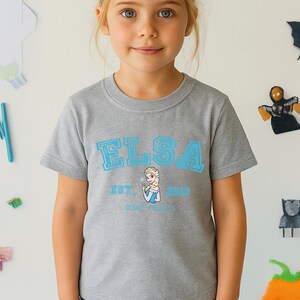 May include: A gray t-shirt featuring the name "ELSA" in light blue block letters, with an image of Elsa and the text "EST. 2013 DISNEY FROZEN". The shirt is made of a soft material and is suitable for children.