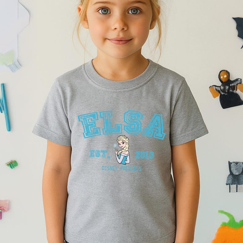 May include: A gray t-shirt featuring the name "ELSA" in light blue block letters, with an image of Elsa and the text "EST. 2013 DISNEY FROZEN". The shirt is made of a soft material and is suitable for children.
